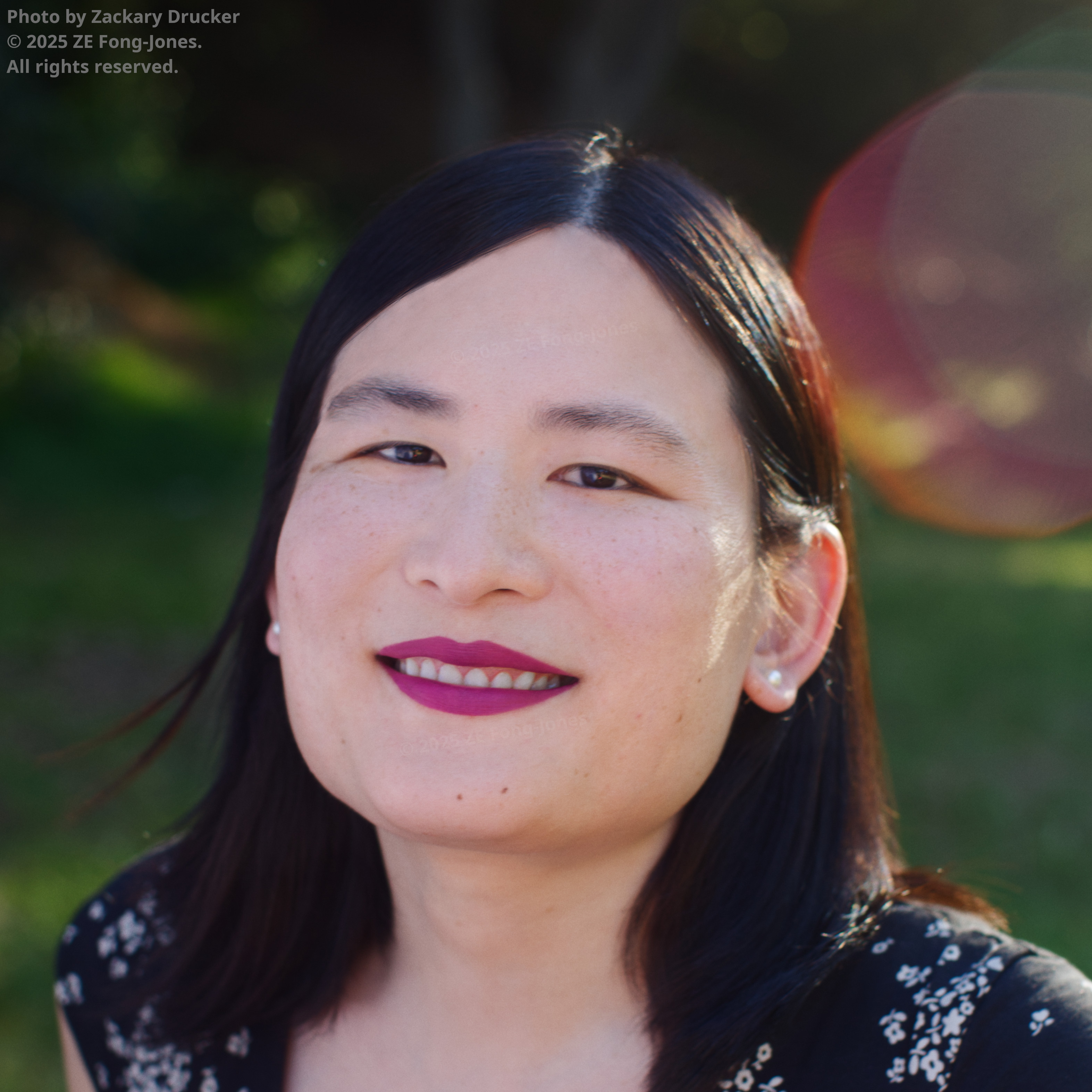 Liz Fong-Jones smiling outdoors, with long black hair, magenta lipstick, and pearl stud earrings, wearing a black floral top